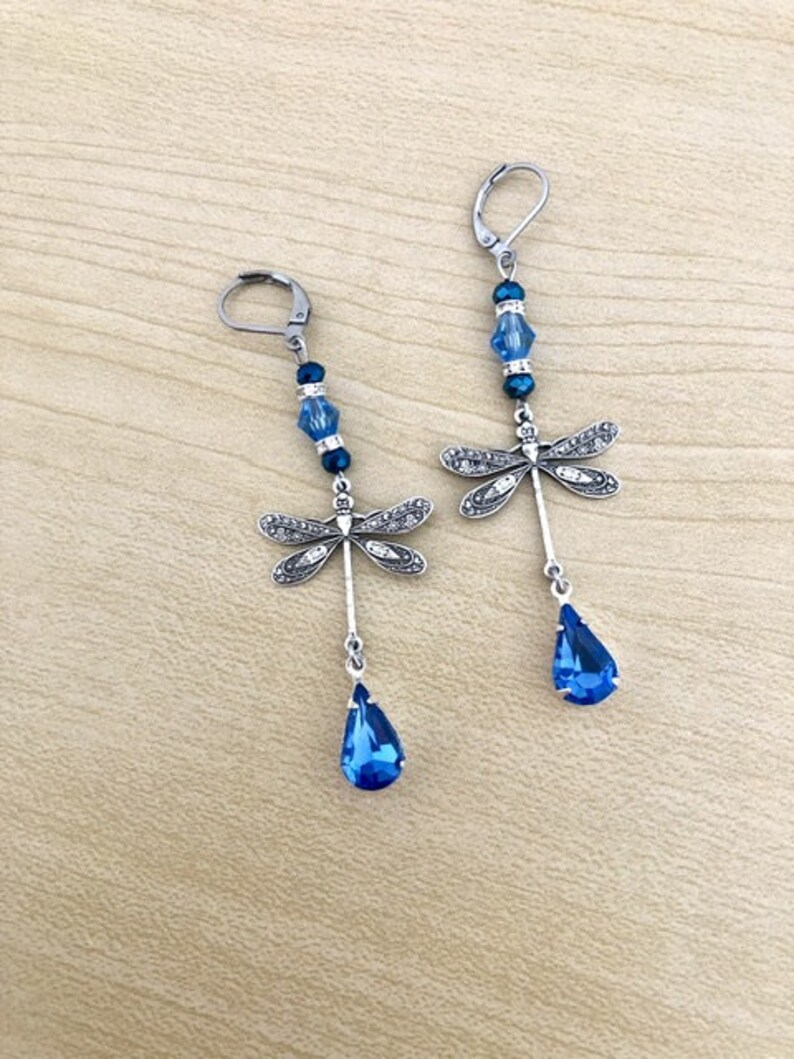 Op de afbeelding: Een paar oorhangers met een zilveren libel-bedel met heldere strass steentjes en blauwe kristallen kralen. De oorhangers hebben een traanvormige blauwe kristal aan de onderkant.