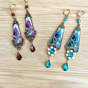 Può includere: Due paia di orecchini pendenti. Un paio presenta un design di iris viola con un pendente a forma di goccia. L'altro paio ha un design di iris blu con un fiore e un pendente a goccia. Entrambi i paia hanno accenti color oro.
