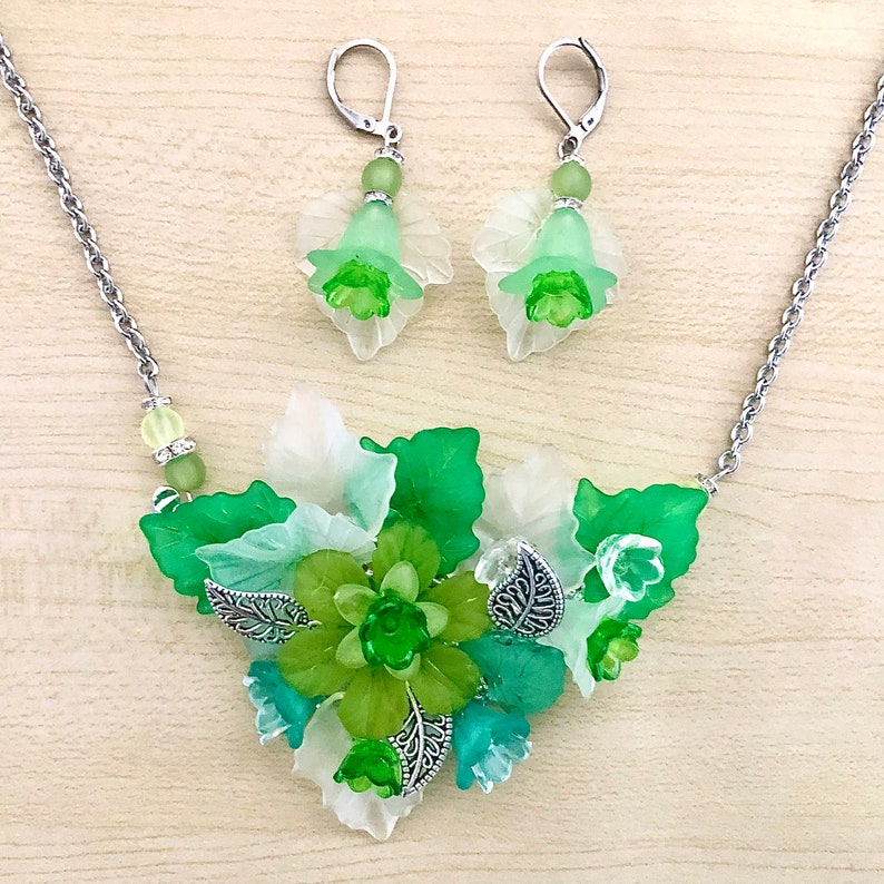 May include: A green and white floral necklace and earrings set with silver accents. The necklace features a cluster of green and white flowers with silver leaves. The earrings are dangle earrings with a single green flower.
