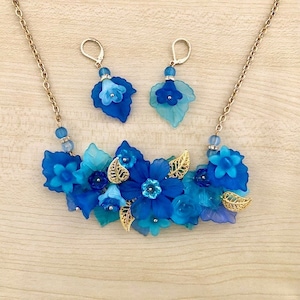 Pu&ograve; includere: Un set di collana e orecchini floreali blu e oro. La collana presenta un gruppo di fiori e foglie blu, con accenti dorati. Gli orecchini sono semplici, con un solo fiore blu.
