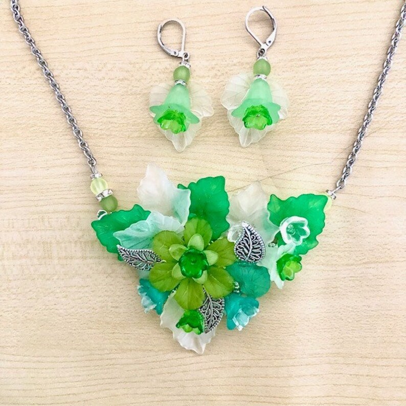 Pu&ograve; includere: Una collana a catena d'argento con un ciondolo floreale verde e bianco. Il ciondolo presenta un gruppo di foglie e fiori in tonalit&agrave; di verde e bianco. La collana include anche un paio di orecchini abbinati con motivi di fiori verdi e bianchi.