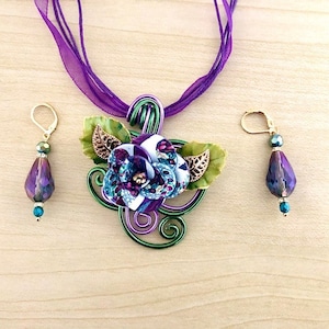 Peut inclure: Un collier avec un pendentif en forme de fleur en fil de fer violet et vert, assorti à des boucles d'oreilles. La fleur est en tissu et a une finition scintillante. Les boucles d'oreilles sont en forme de goutte et ont une finition violette irisée.