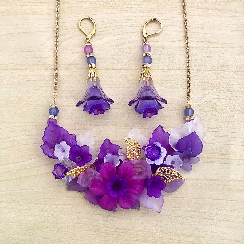 May include: Purple and white flower necklace and earrings set. The necklace features a cluster of purple and white flowers with gold accents. The earrings are dangle earrings with purple flowers.