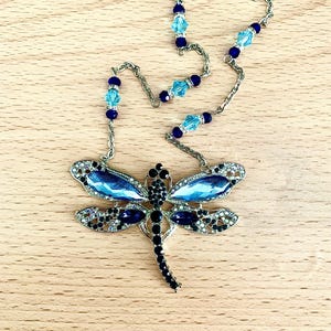 Op de afbeelding: Een zilveren ketting met blauwe en zwarte kristallen kralen en een libellenhanger met blauwe en zwarte kristallen accenten.