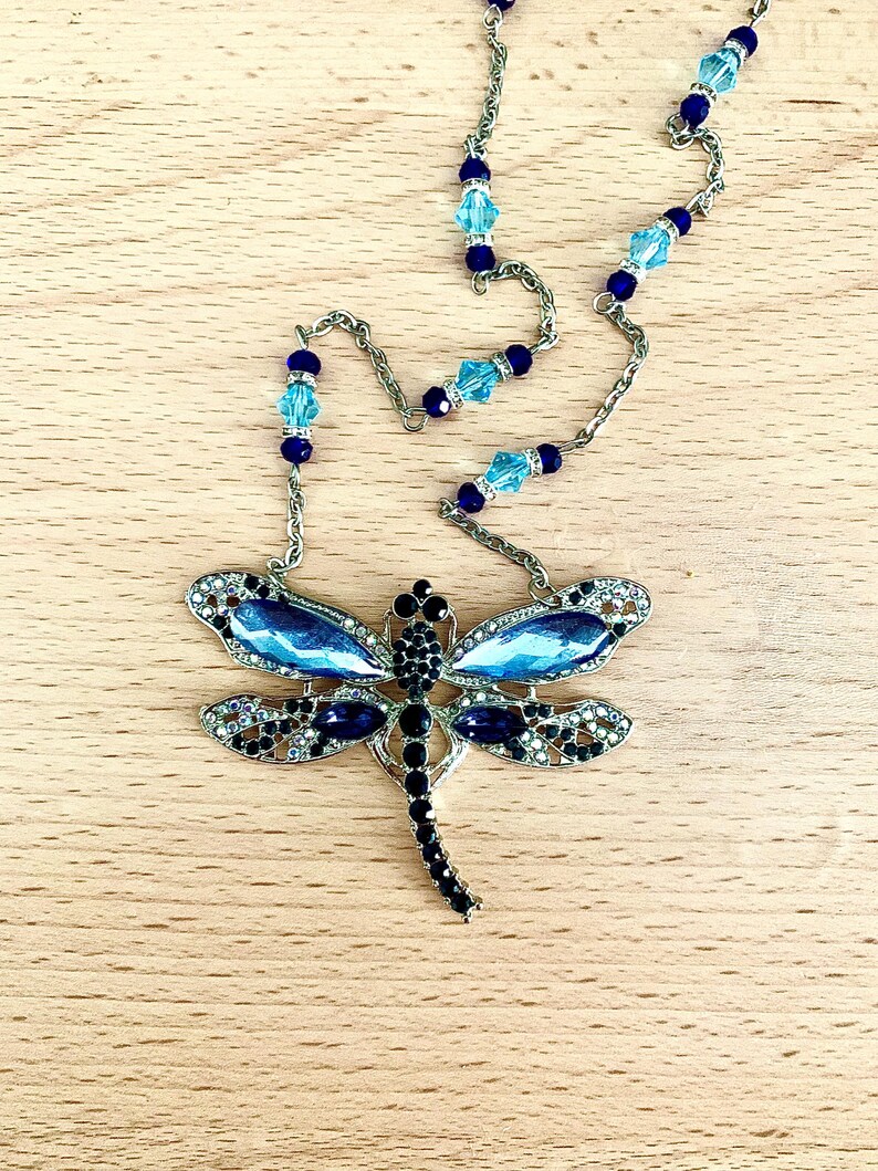Op de afbeelding: Een zilveren ketting met blauwe en zwarte kristallen kralen en een libellenhanger met blauwe en zwarte kristallen accenten.