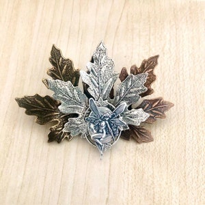 Peut inclure: Un charme de f&eacute;e argent&eacute; avec des ailes, assis dans un cercle de feuilles. Les feuilles sont en m&eacute;tal et sont dans des tons de bronze et d'argent.