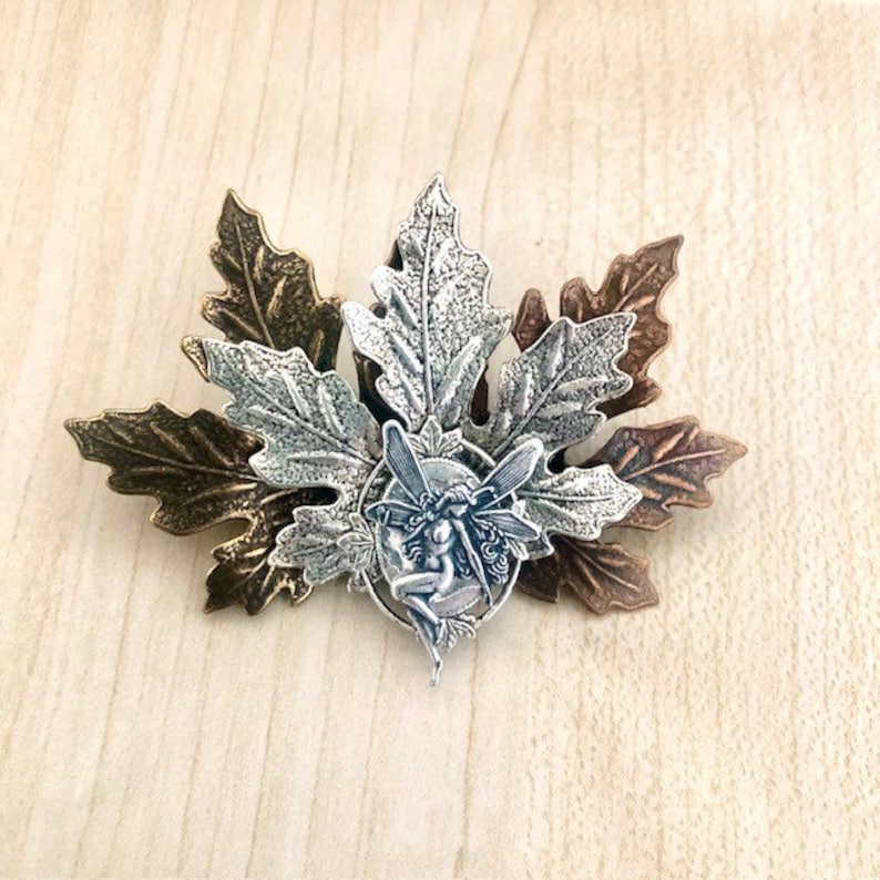 Peut inclure: Un charme de f&eacute;e argent&eacute; avec des ailes, assis dans un cercle de feuilles. Les feuilles sont en m&eacute;tal et sont dans des tons de bronze et d'argent.