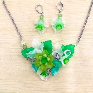 May include: A green and white floral necklace and earrings set. The necklace features a cluster of green and white flowers with silver leaves. The earrings are simple green and white flowers with silver hooks.