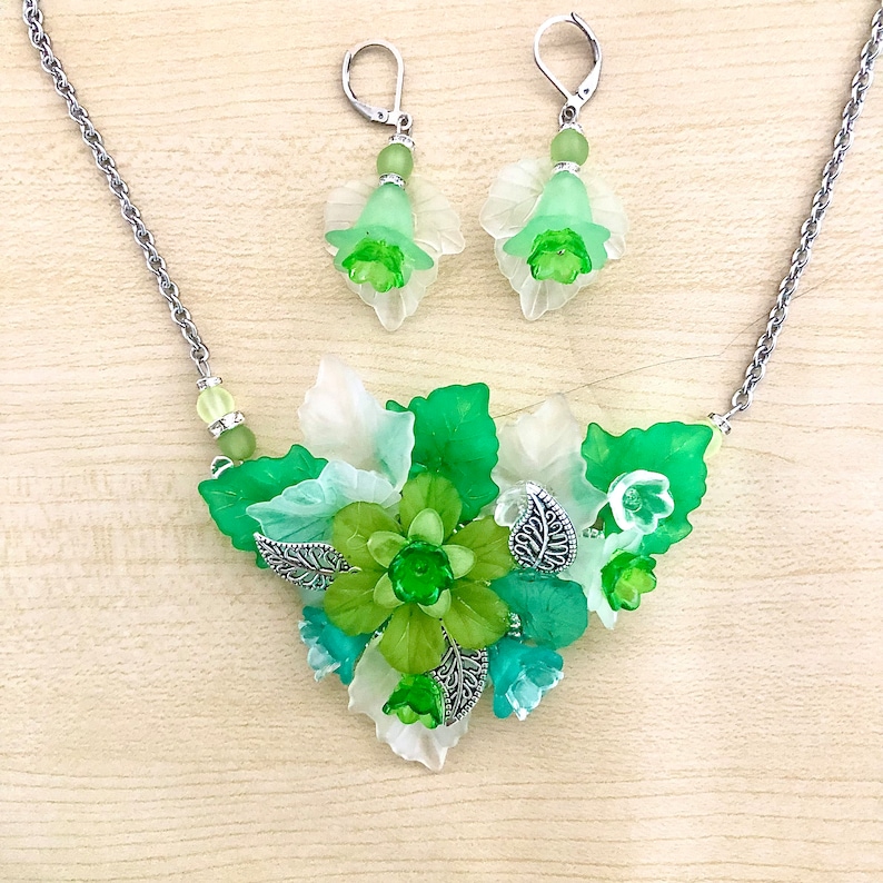 May include: A green and white floral necklace and earrings set. The necklace features a cluster of green and white flowers with silver leaves. The earrings are simple green and white flowers with silver hooks.