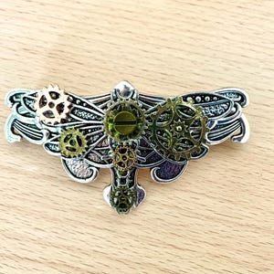 Pode incluir: Um prendedor de cabelo de metal prateado e verde em forma de libélula com detalhes intrincados de engrenagens. As asas da libélula estão abertas e as engrenagens são visíveis no corpo e nas asas.