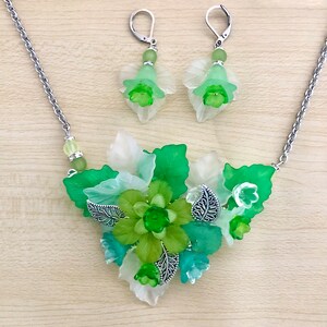 May include: A green and white floral necklace and matching earrings. The necklace features a cluster of green and white flowers with silver accents. The earrings are simple and feature a single green flower.