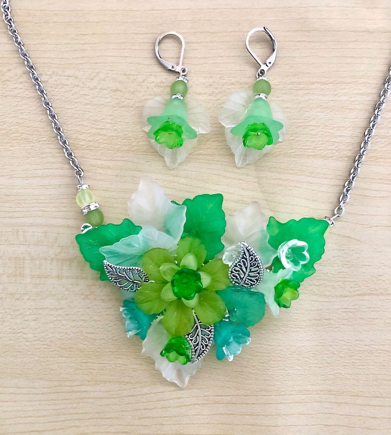 May include: A green and white floral necklace and matching earrings. The necklace features a cluster of green and white flowers with silver accents. The earrings are simple and feature a single green flower.