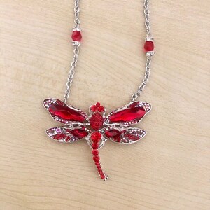 Op de afbeelding: Een zilveren ketting met een rode, met edelstenen bezette libel hanger. De libel heeft een gedetailleerd ontwerp met rode en heldere kristallen.