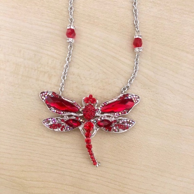 Op de afbeelding: Een zilveren ketting met een rode, met edelstenen bezette libel hanger. De libel heeft een gedetailleerd ontwerp met rode en heldere kristallen.
