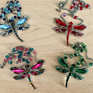 Op de afbeelding: Vier kettingen met libellen hangers. Elke ketting heeft een libellen hanger in een andere kleur met kristal accenten. De libellen zijn blauw, rood, roze en groen.