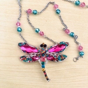 Op de afbeelding: Een zilveren ketting met een roze en veelkleurige libel hanger. De libel is versierd met strass steentjes en heeft een lange, parelbezet staart.