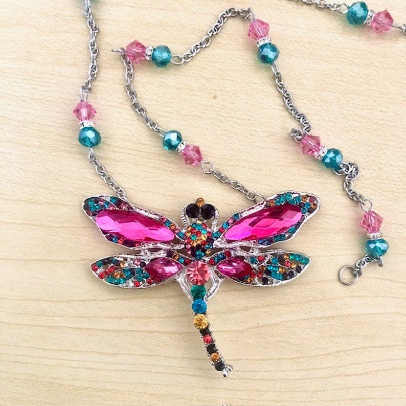 Op de afbeelding: Een zilveren ketting met een roze en veelkleurige libel hanger. De libel is versierd met strass steentjes en heeft een lange, parelbezet staart.