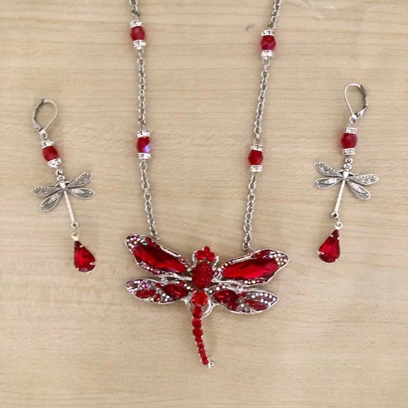 Op de afbeelding: Een rode kristallen libellen hangerketting en bijpassende oorbellen. De libel is versierd met zilveren accenten en rode kristallen. De ketting heeft een zilveren ketting met rode kristallen kralen.