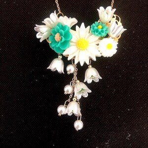 Op de afbeelding: Een witte en groene bloemenketting met een gouden ketting. De ketting heeft een cluster van witte bloemen met groene centra en een hangende cluster van witte, klokvormige bloemen met parel accenten.