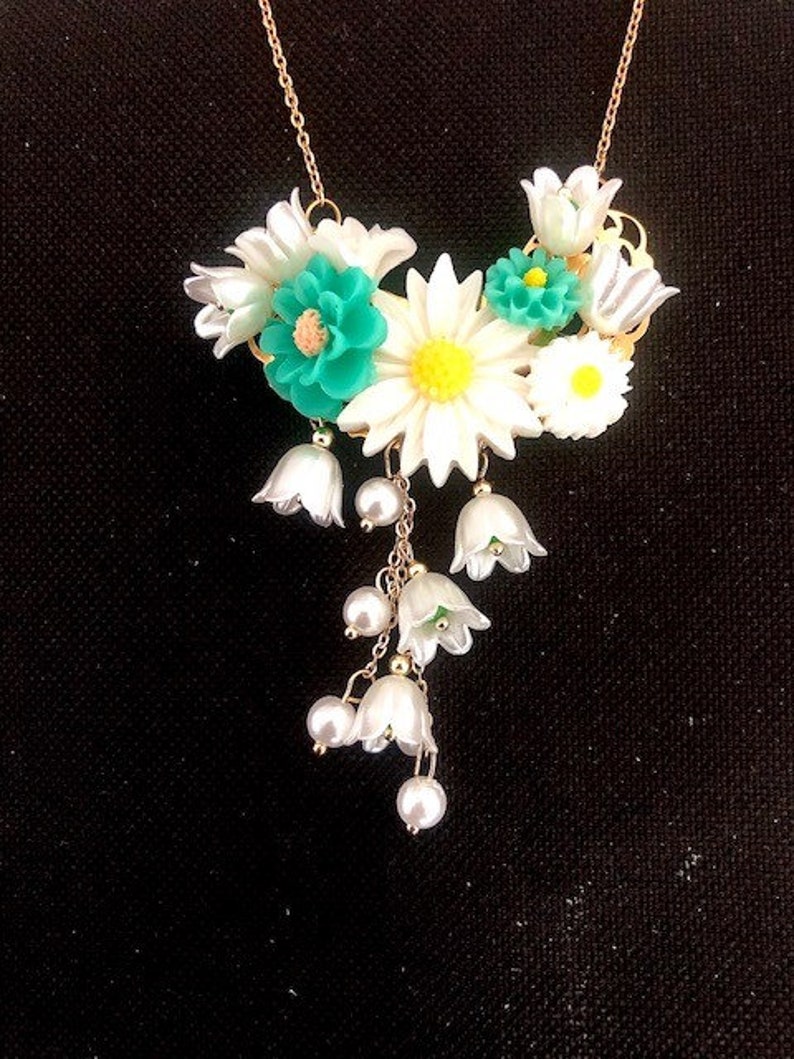 Op de afbeelding: Een witte en groene bloemenketting met een gouden ketting. De ketting heeft een cluster van witte bloemen met groene centra en een hangende cluster van witte, klokvormige bloemen met parel accenten.