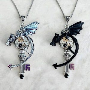 Peut inclure: Deux colliers avec des pendentifs en forme de dragon enroulés autour d'un mécanisme d'horlogerie et d'une base en forme de clé verticale. Un dragon est argenté avec des reflets métalliques, l'autre est noir uni. Les deux pendentifs sont sur des chaînes.