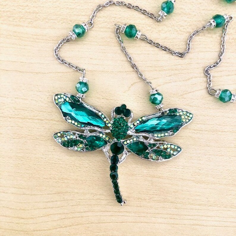 Op de afbeelding: Een zilveren ketting met een grote groene libel hanger. De libel is gemaakt van strass en heeft een lange, slanke staart.