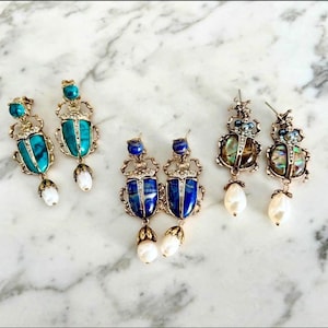May include: Three pairs of gold-tone beetle-shaped earrings. The first pair features turquoise-colored stones, the second pair features deep blue stones, and the third pair features iridescent abalone shells. Each earring has a dangling white pearl.