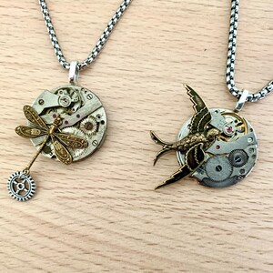 May include: Two silver necklaces with unique pendants. One pendant features a dragonfly with a gear charm, the other a bird with a watch face.