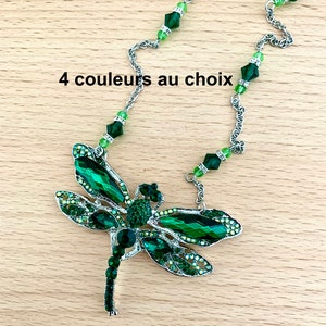 Op de afbeelding: Een zilveren ketting met een groene libel hanger. De libel is gemaakt van metaal en heeft groene kristallen accenten. De ketting is versierd met groene kristallen kralen. "4 couleurs au choix" staat in zwarte tekst op een houten oppervlak.