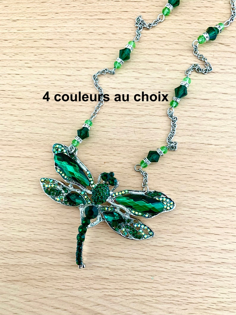 Op de afbeelding: Een zilveren ketting met een groene libel hanger. De libel is gemaakt van metaal en heeft groene kristallen accenten. De ketting is versierd met groene kristallen kralen. "4 couleurs au choix" staat in zwarte tekst op een houten oppervlak.