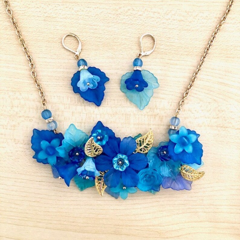 Pu&ograve; includere: Un set di collana e orecchini floreali blu e oro. La collana presenta un gruppo di fiori e foglie blu, con accenti dorati. Gli orecchini sono semplici, con un solo fiore blu su ciascuno.