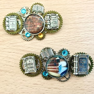 Könnte beinhalten: Zwei goldfarbene Broschen im Buch-Design mit blauen und goldenen Akzenten. Die Broschen haben eine runde Glaskuppel mit einem Bild von Büchern darin. Eine Brosche trägt den Text "Once Upon A Time" und die andere den Text "A True Story".