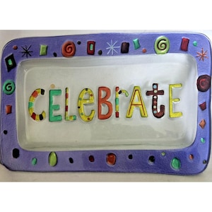 Puede incluir: Una fuente rectangular de vidrio con un borde morado decorado con formas coloridas. La palabra "CELEBRATE" está escrita en letras coloridas en el centro de la fuente.