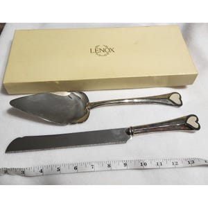 Può includere: Un set di coltello e paletta per torta argentati con dettagli a forma di cuore sui manici, presentato con una scatola Lenox color crema. Il coltello ha un bordo seghettato. Un metro a nastro è posto vicino al set.