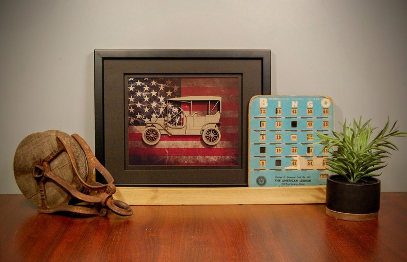 May include: A framed print of a vintage car in front of an American flag. The print is on a wooden shelf with a wooden spool, a bingo card, and a potted plant.
