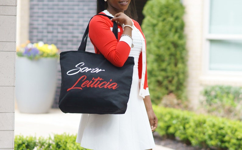 May include: A black tote bag with white and red lettering that says "Soror Leticia".