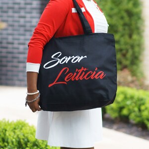 May include: A black tote bag with white and red lettering that says "Soror Leticia".