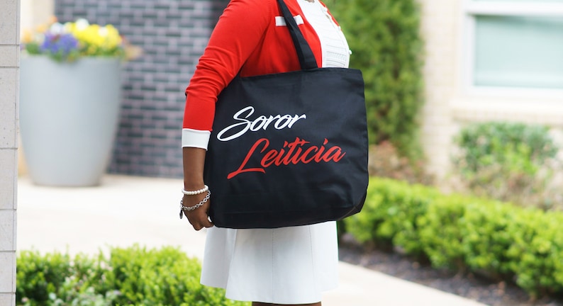 May include: A black tote bag with white and red lettering that says "Soror Leticia".