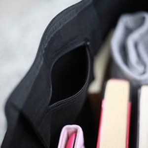 May include: A black zippered tote bag with a zippered pocket on the front. The bag is open and filled with books and a gray knitted scarf.