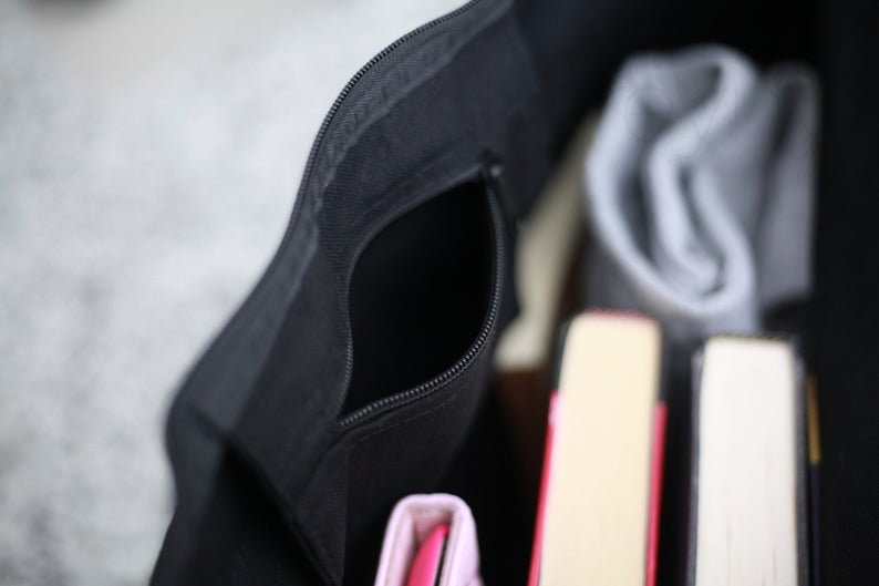 May include: A black zippered tote bag with a zippered pocket on the front. The bag is open and filled with books and a gray knitted scarf.