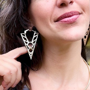 May include: A woman with dark brown hair wearing a grey tank top and a gold choker necklace. She is wearing large gold geometric earrings. The earrings are shaped like a triangle with a circle in the centre.