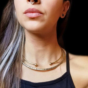 May include: A woman with dark hair and a silver streak in her hair is wearing a black tank top and a gold choker necklace. She has a nose ring and a small earring in her right ear.