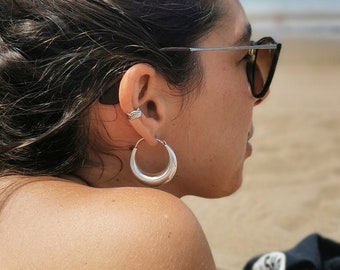 May include: A woman with long brown hair pulled back in a bun, wearing sunglasses and hoop earrings, lies on a beach towel on a sandy beach.