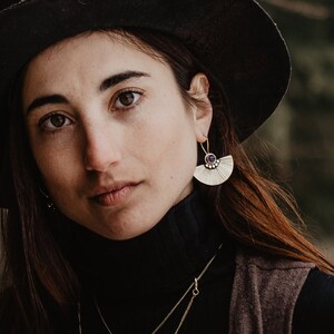May include: A person wearing a black hat, black turtleneck, and a brown vest. They are wearing gold necklaces and earrings. The background is blurred with green and brown tones.
