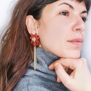 May include: A woman wearing a gray turtleneck sweater with long red and gold dangle earrings. The earrings feature a red, intricate design with a gold chain.