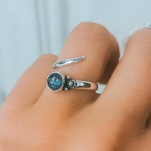 Op de afbeelding: Een zilveren ring met een blauwe edelsteen en een gedraaide band. De ring zit aan een vinger.