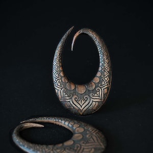 May include: Two wooden ear gauges with intricate carved designs. The gauges are a dark brown colour with a copper finish. The designs are symmetrical and feature a repeating pattern of circles and swirls.