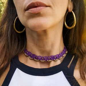 Peut inclure: Une femme portant un débardeur blanc avec une bordure noire et un collier ras du cou en perles violettes et dorées. Elle porte de grandes boucles d'oreilles créoles dorées.