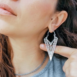 May include: A woman with long brown hair is wearing a silver geometric hoop earring. The earring is shaped like a triangle with intricate details.