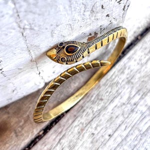 Könnte beinhalten: Ein goldfarbenes Armband in Schlangenform. Der Schlangenkopf hat einen dunkelroten Edelstein und detaillierte Schuppenmuster. Das Armband ist um ein Stück verwittertes Holz gewickelt.